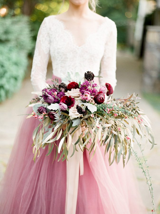 In questa foto una sposa con abito in pizzo bianco e gonna in tulle rosa tiene tra le mani un bouquet a braccio con foglie, rose rosa e rosse, tulipani rosa e ranuncoli bordeaux