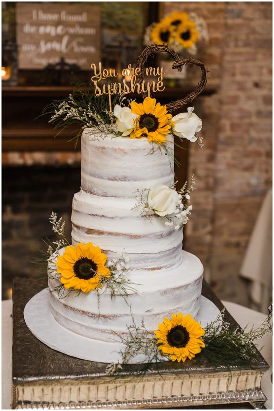 In questa foto una naked cake per un matrimonio a tema girasoli adagiati sui tre piani insieme a boccioli di rosa bianchi e a fiori velo di sposa. Il cake topper dice "You are my sunshine" cioè "Sei la mia luce del sole"