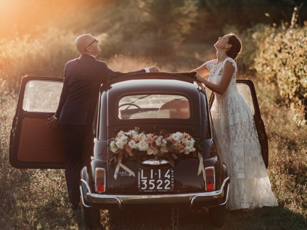 In questa foto di Studio fotografico Righi gli sposi posano divertiti davanti ad una Cinquecento antica   