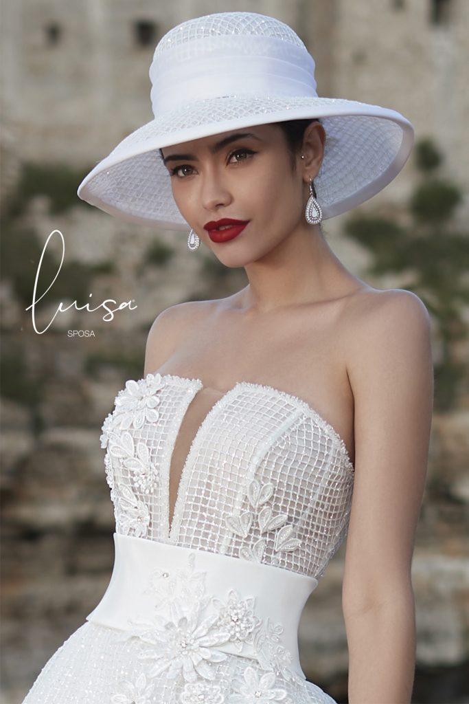 In questa foto una modella indossa un abito con scollo a V e ricami 3D sul corpetto e sulla cintura e un cappello da sposa modello Borsalino 