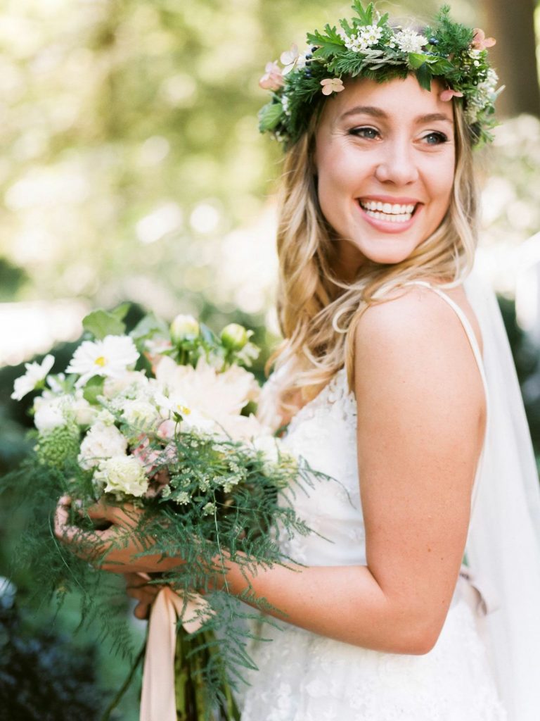 In questa foto una sposa indossa una corona di foglie, fiori bianchi e rosa e tiene tra le mani un bouquet a braccio abbinato. La sposa è girata e sorride