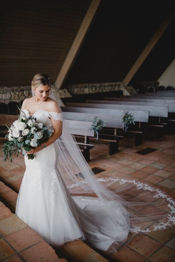 In questa foto una sposa all'interno di una cappella che guarda in basso mentre stringe il suo bouquet a braccio di fiori bianchi ed eucalipto. La sposa indossa un abito a mezza sirena e un velo Chiesa con pizzo rebrodè