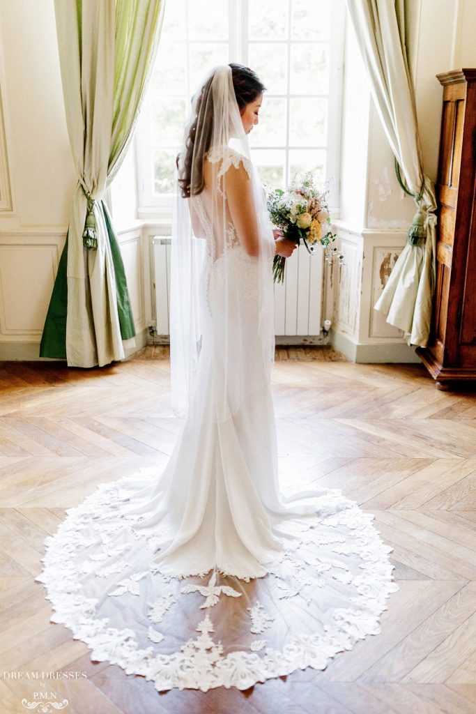 In questa foto una sposa è ritratta nella stanza di un antico palazzo mentre guarda il suo bouquet e indossa un velo Floor con bordo in pizzo Rebrodè  abbinato ad un abito a sirena con scollatura sulla schiena effetto tattoo