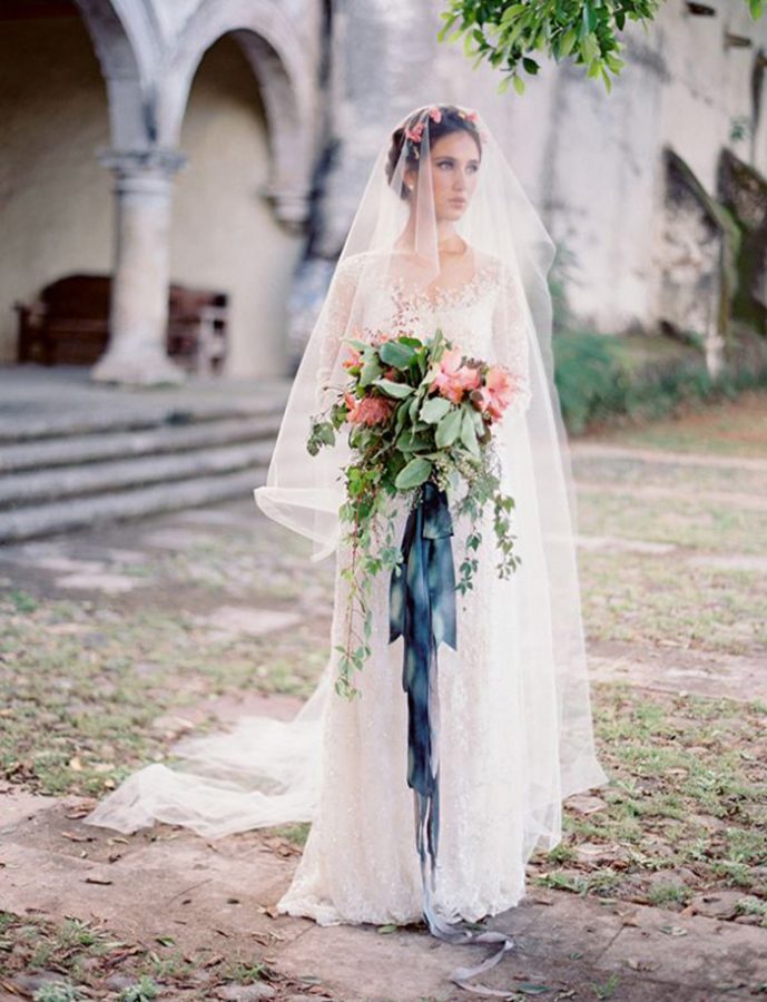 In questa foto una sposa con corona di fiori e velo con calata porta tra le mani un velo a braccio di fogliee ninfee raccolte da un nastro blu