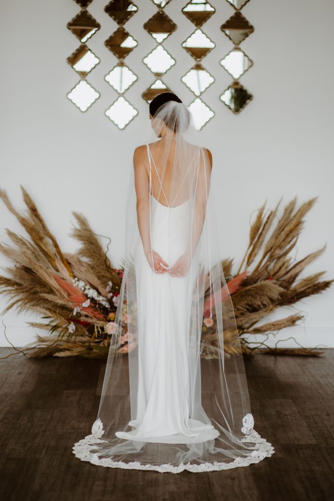 In questa foto una modella ritratta di spalle indossa un velo a cappella  in tulle di seta con bordo in pizzo rebrodè fissato su uno chignon. La sposa indossa un abito a sirena morbido in seta con scollatura posteriore