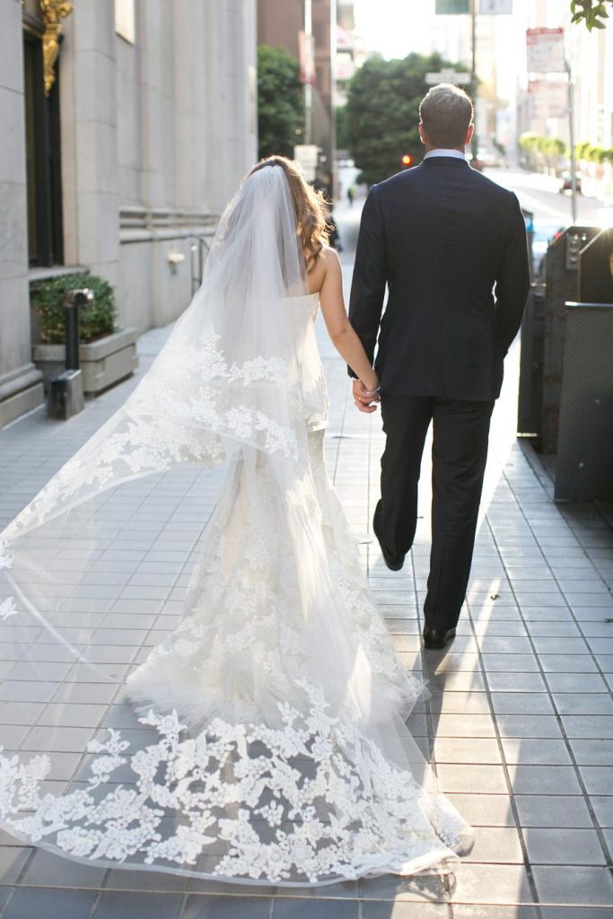 In questa foto due sposi che camminano per la strada visti di spalle. La sposa indossa un velo a cappella con pizzo rebrodè abbinato all'abito a mezza sirena