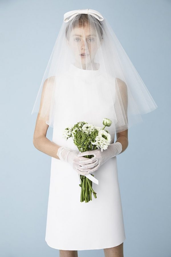 In questa foto una modella indossa un velo da sposa a petalo in tulle di seta con fiocco sul capo che le copre completamente il viso. Il velo è abbinato a un tubino e a dei guanti e porta tra le mani un bouquet di fiori di campo bianchi