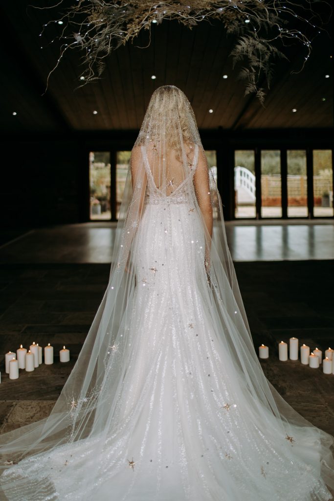 In questa foto una sposa viene ripresa   di spalle in una sala e davanti delle candele accese poggiate in terra con il suo velo di brillantini dorati in primo piano