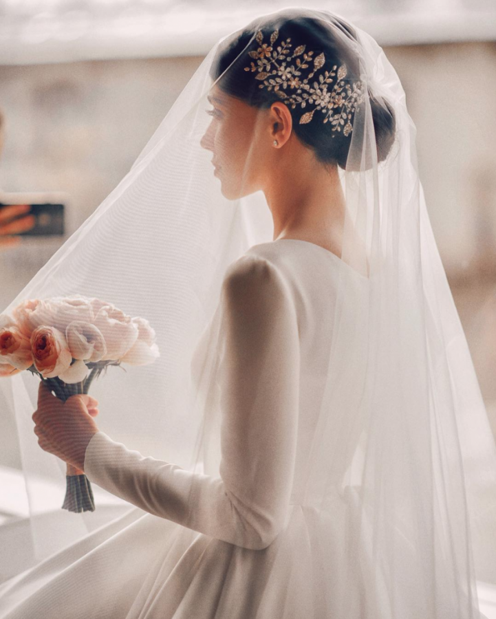 In questa foto una sposa si dirige all'altare sotto al suo velo di tulle e tiene nella mano sinistra un bouquet di peonie e rosa e bianche. La sposa indossa un abito ampio con maniche lunghe semitrasparenti
