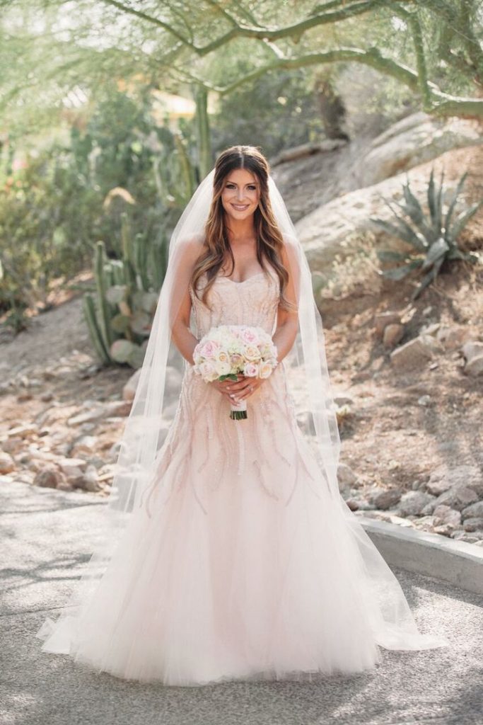 In questa foto una sposa con abito rosa cipria e ramage di perline posa in un giardino di succulente, con il suo velo medio di tulle a taglio vivo e tenendo tra le mani un bouquet di rose bianche e rosa