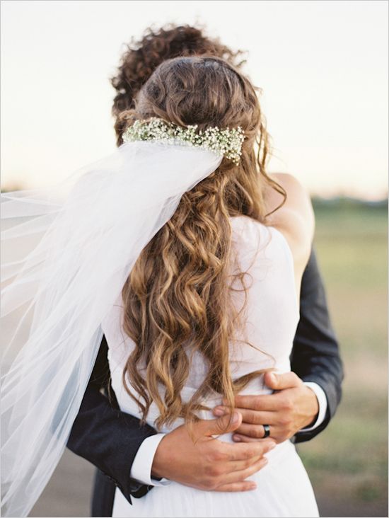 In questa foto due sposi che si abbracciano in un prato. La sposa è di spalle e mostra il velo che ondeggia al velo attaccato sulla nuca ai capelli semiraccolti intrecciati con dei fiori velo da sposa