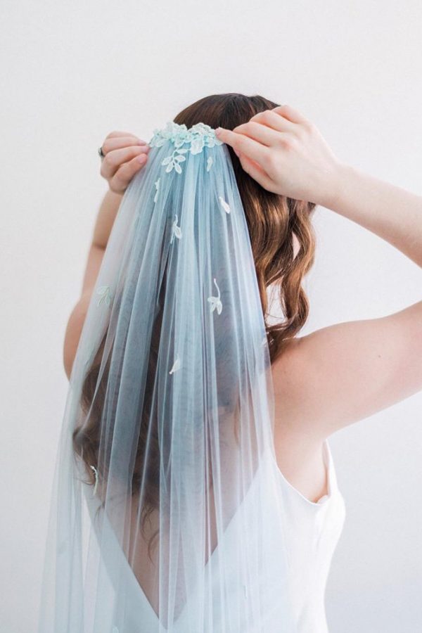 In questa foto una sposa aggiusta sulla nuca il suo velo colore azzurro con ricami di fiori in 3D