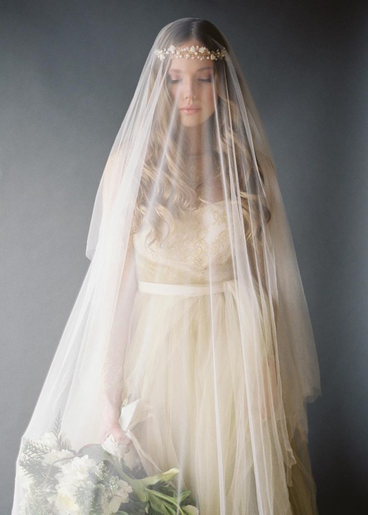 In questa foto una sposa ad occhi chiusi indossa un abito da sposa colore champagne e velo abbinato tenendo verso il basso il bouquet con la manco destra