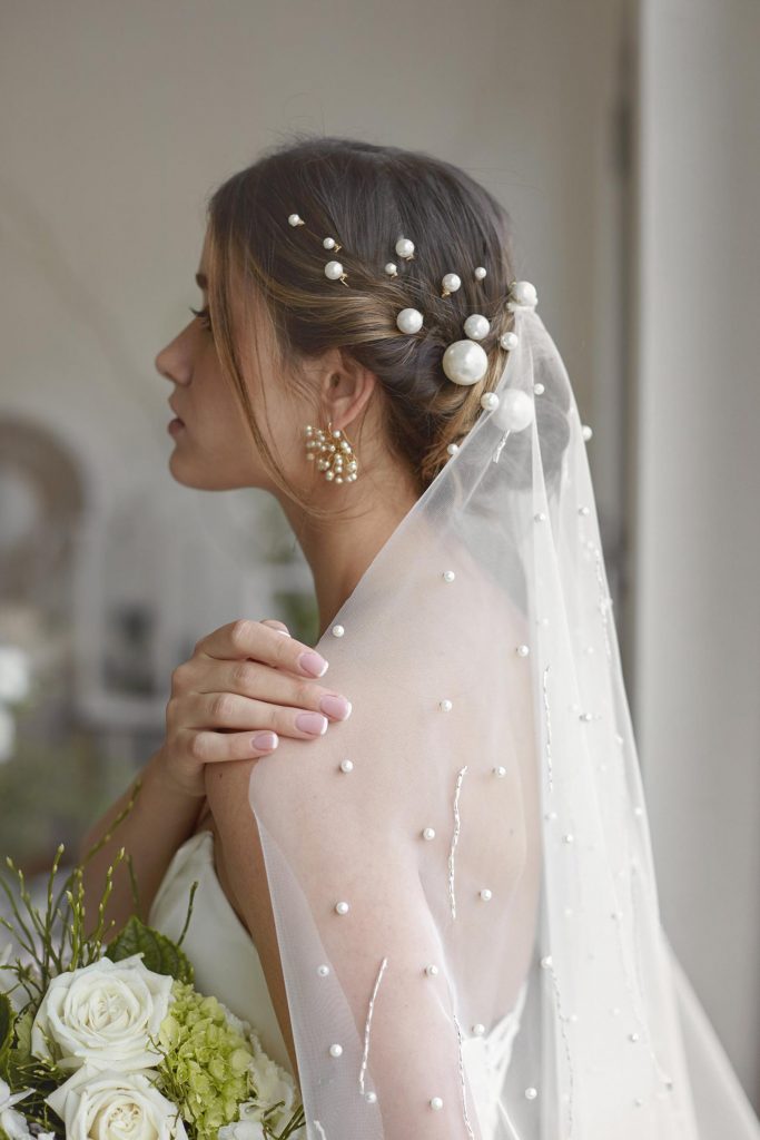 In questa foto una modella posa di profilo con un velo decorate con perline e forcine di perle grandi e piccole che si intrecciano ai capelli