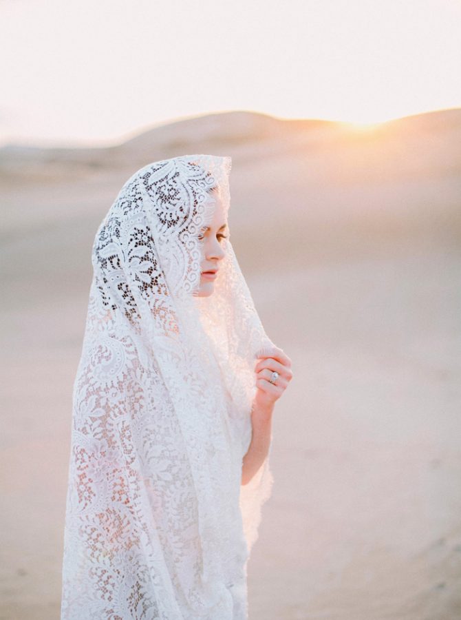 In questa foto una sposa è ripresa del deserto con il suo velo interamente in pizzo rebrodato portato a mantiglia