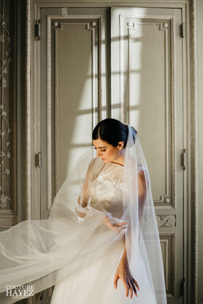 In questa foto una sposa ripresa di tre quarti avvicina con la mano destra il suo velo da sposa in seta Couture Hayez al braccio sinistro