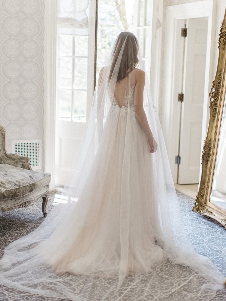 In questa foto una sposa ripresa di spalle in una stanza di hotel mentre indossa il suo velo da sposa di tulle