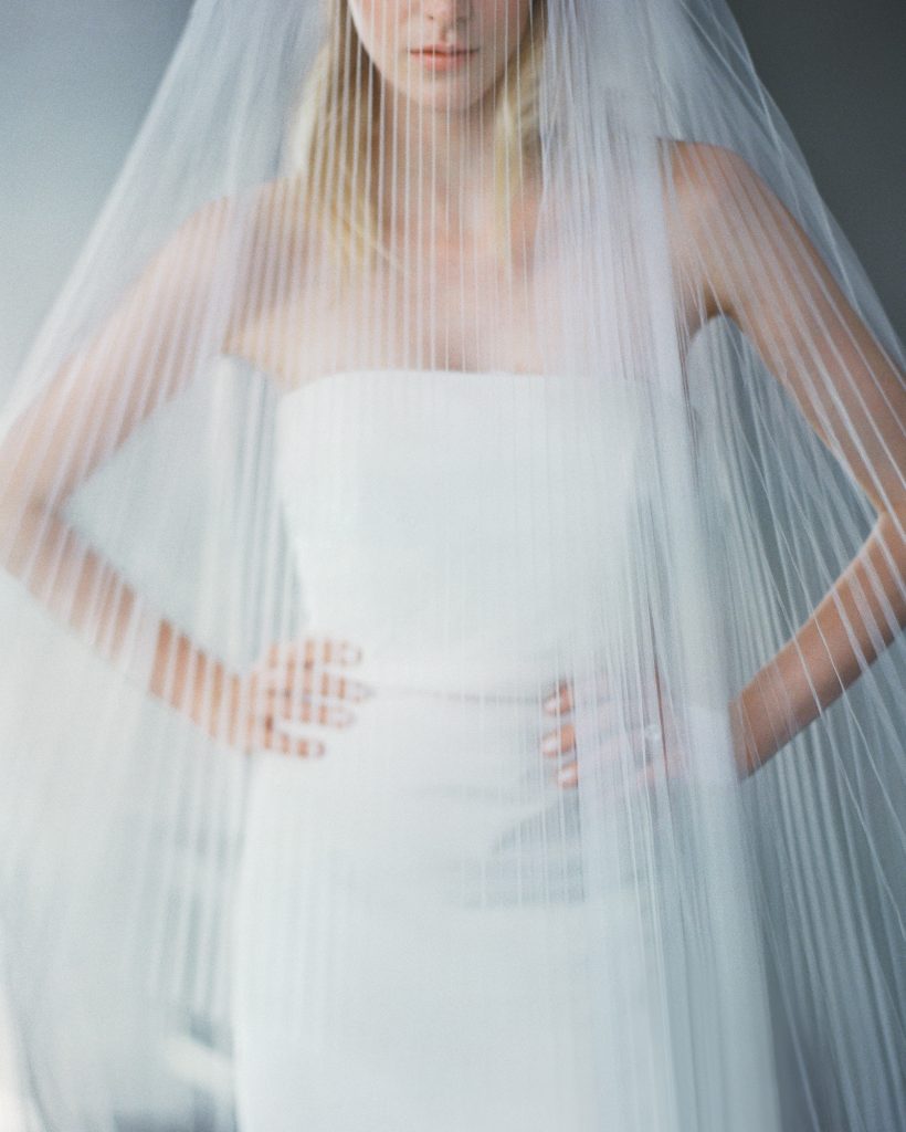 In questa foto una sposa sotto il suo velo plissettato posa con le braccia poggiate sui fianchi. La sposa è ripresa dal naso in giù