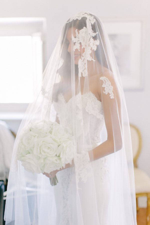 In questa foto una sposa viene ripresa sotto il suo velo con calata mentre sorride e tiene tra le sue mani un bouquet di rose bianche.