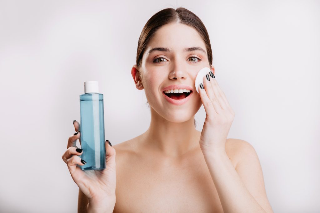 In questa foto una ragazza sorridente a bocca aperta strofina con la mano sinistra un dischetto di cotone mentre nell'altra mano tiene una bottiglietta azzurra di struccante 