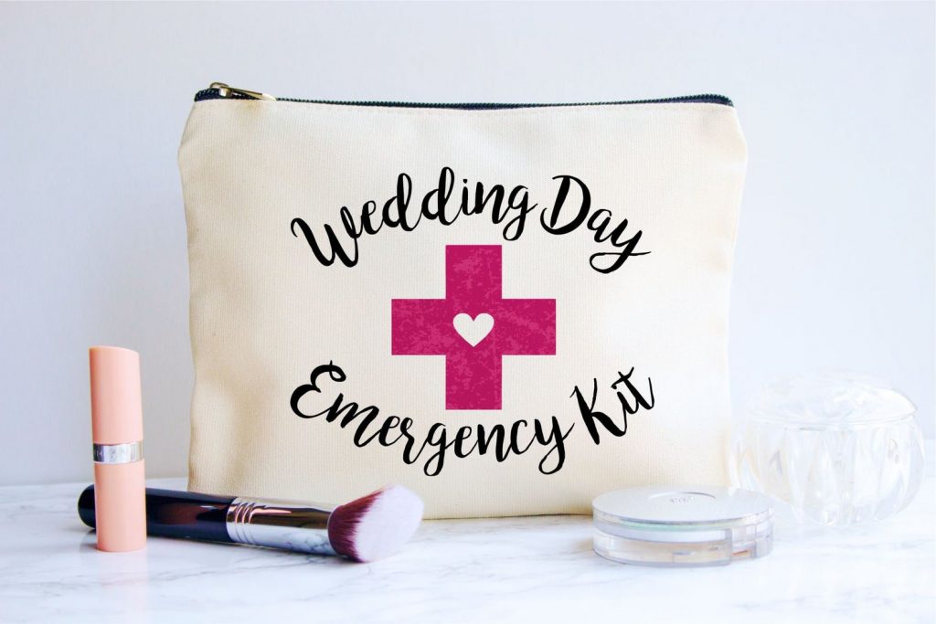 In questa foto una borsetta bianca rettangolare con scritto Wedding Day Emergency kit. Davanti sono poggiati un rossetto, un pennello e una cipria