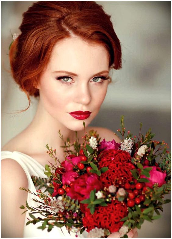 In questa foto una modella con capelli rossi e occhi verdi tiene tra le mani un bouquet di fiori rossi e fuxia. Indossa un trucco che mette in evidenza le labbra con un rossetto colore vino semilucido. Completa il look l'eyeliner nero