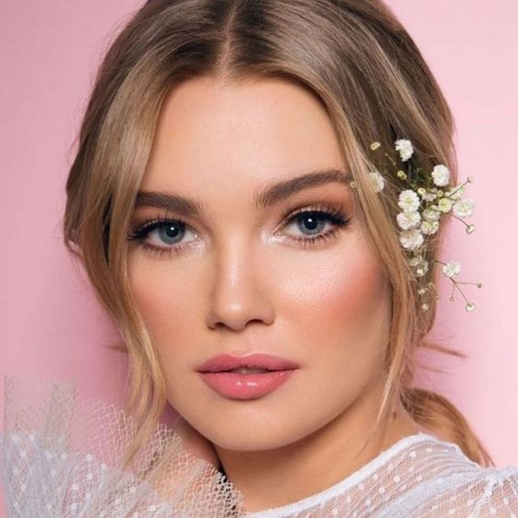 In questa foto un modella bionda con un raccolto morbido completato da fiori velo da sposa è truccata con un blush nei toni del rosa e rossetto lucido abbinato 