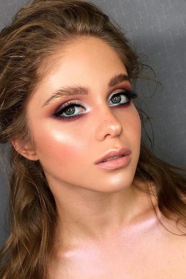 In questa foto un modella con capelli colore biondo scuro indossa un trucco luminoso realizzato con ombretti shimmer lilla, rosa e colore  argento, matita azzurra nella rima interna dell'occhio e illuminante su zigomi, mento e naso