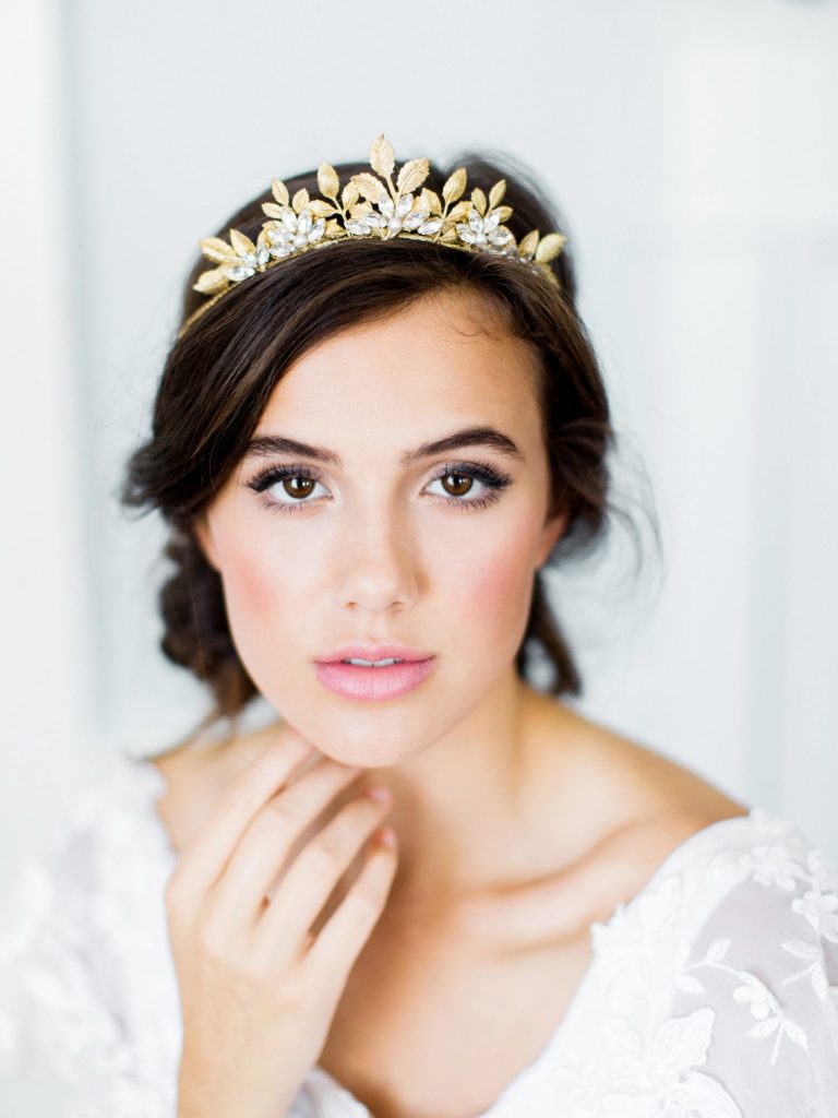 In questa foto una modella che posa con una mano poggiata sotto il mento. Indossa un abito da sposa con ricami floreali e mostra il suo trucco sposa naturale realizzato con un rossetto rosa, un blush abbinato e un eyeliner sfumato. Il look è completato con un acconciatura raccolta con una coroncina di foglie colore oro e strass