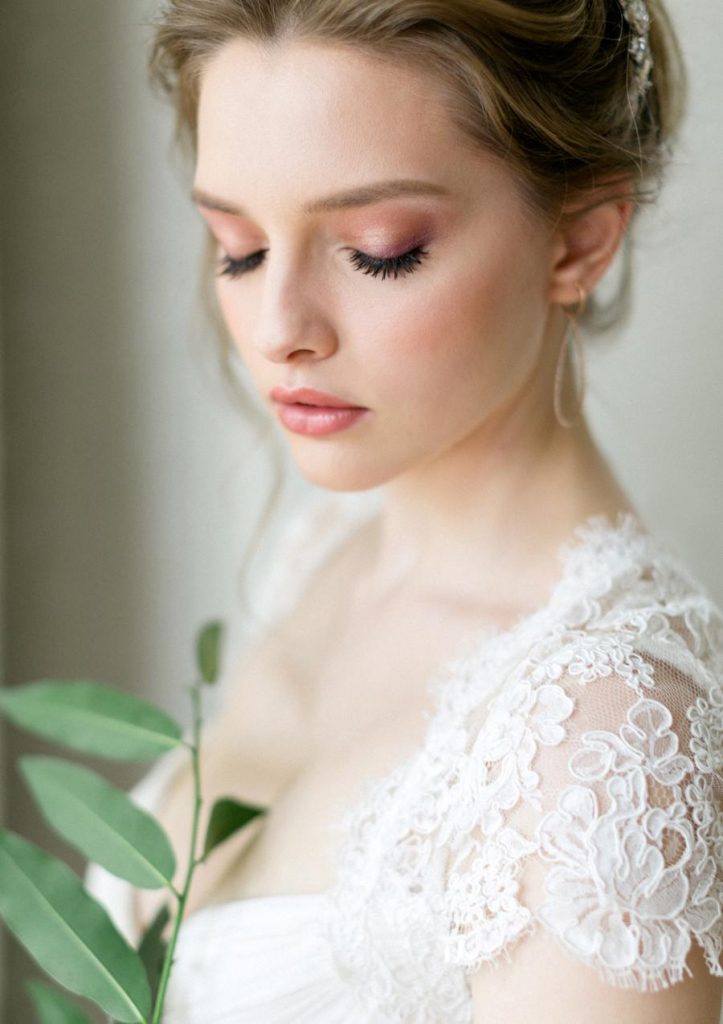 In questa foto una sposa bionda con pelle chiara ritratta ad occhi chiusi con un trucco naturale nei toni del pesca. Indossa un abito da sposa in pizzo rebrodè a maniche corte e tiene tra le mani un rametto di foglie