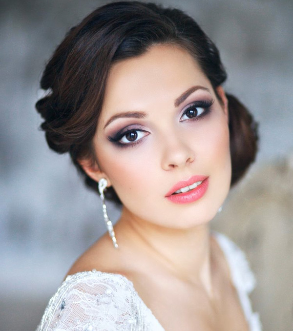 In questa foto una sposa con capelli raccolti indossa uno smokey eyes make up nei toni del viola e del lilla. Sulle labbra porta un rossetto semilucido colore rosa e sulle guance un blush abbinato
