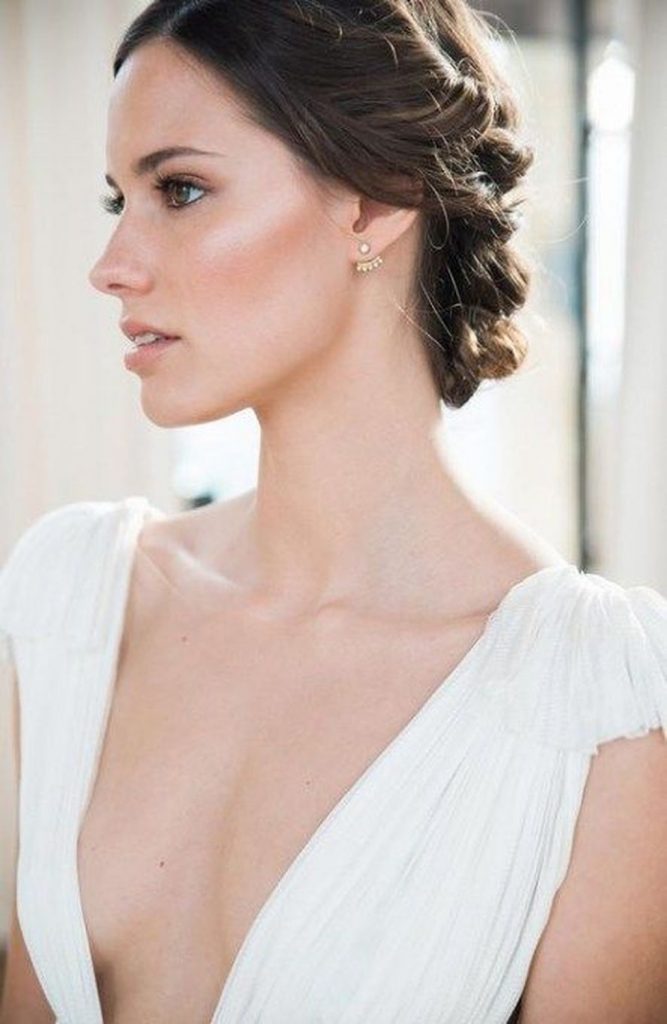 In questa foto una sposa ritratta di profilo mentre guarda a sinistra mostra un piccolo orecchino a chiodo. Indossa un abito con una profonda scollatura a V drappeggiata sul seno e sulle spalle. I capelli sono raccolti in uno chignon morbido composto di trecce. 