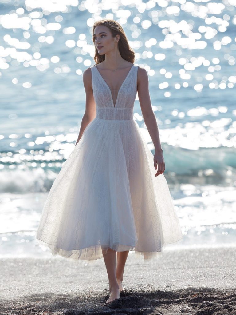 In questa foto una modella sulla spiaggia cammina indossando un abito da sposa midi per matrimonio civile in tulle glitter, scivolato e con scollatura a V firmato Nicole Milano  Collection