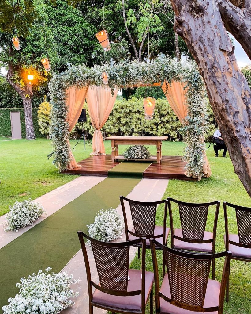 In questa foto un allestimento per un matrimonio civile in giardino al tramonto. Al centro è posto un gazebo di legno decorato con foglie rampicanti e drappi di lino beige. Al centro del baldacchino è presente un tavolino di legno con un cesto di fiori bianchi e foglie. Dal baldacchino parte un tappeto verde e ai lati sono presenti sedie di legno rivestite di tessuto color lavanda. Ai rami degli alberi sono attaccate alcune lanterne con candele accese