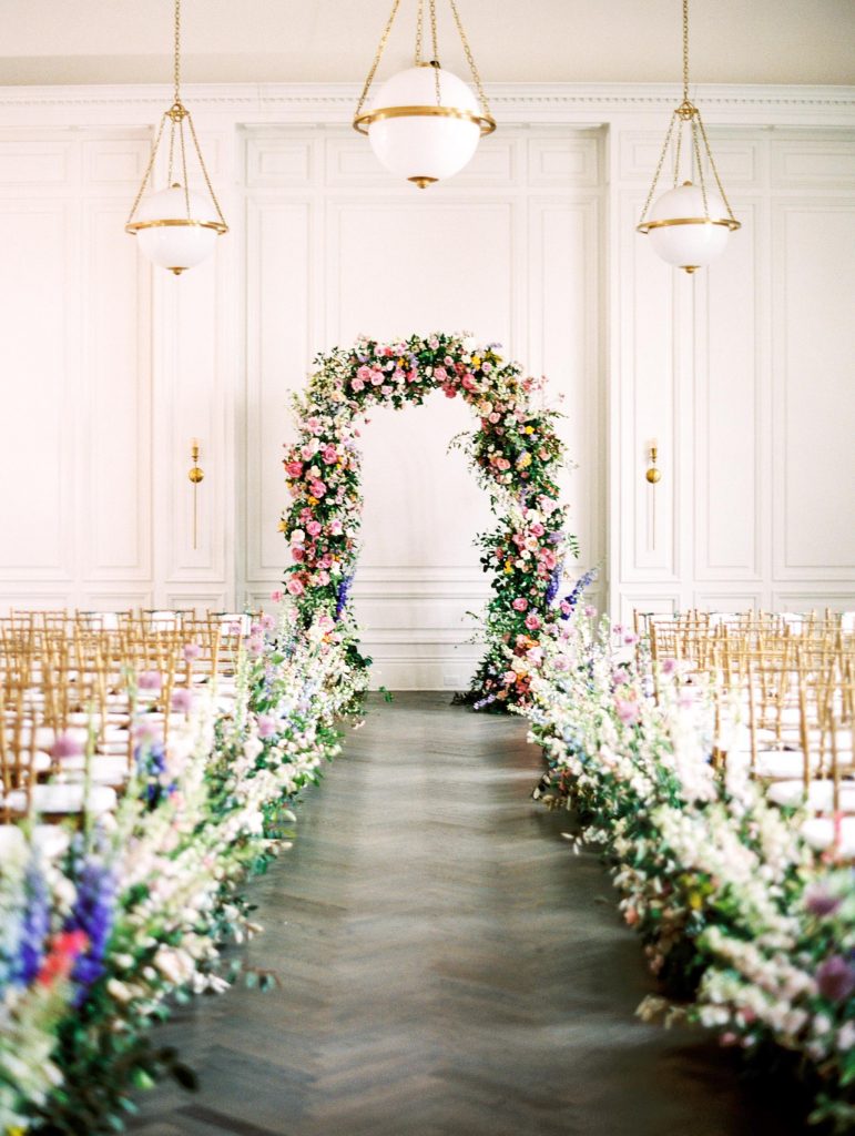 In questa foto l'allestimento di un matrimonio civile nella sala di una villa con sedie colore oro e bianche e composizioni di fiori di campo lungo il corridoio e che ricoprono interamente l'arco dove si celebrerà il rito