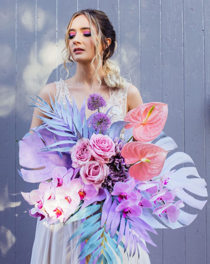 In questa foto una sposa posa con il suo bouquet multicolor dai toni del rosa e dele viola con palme, rose, orchidee e piante grasse