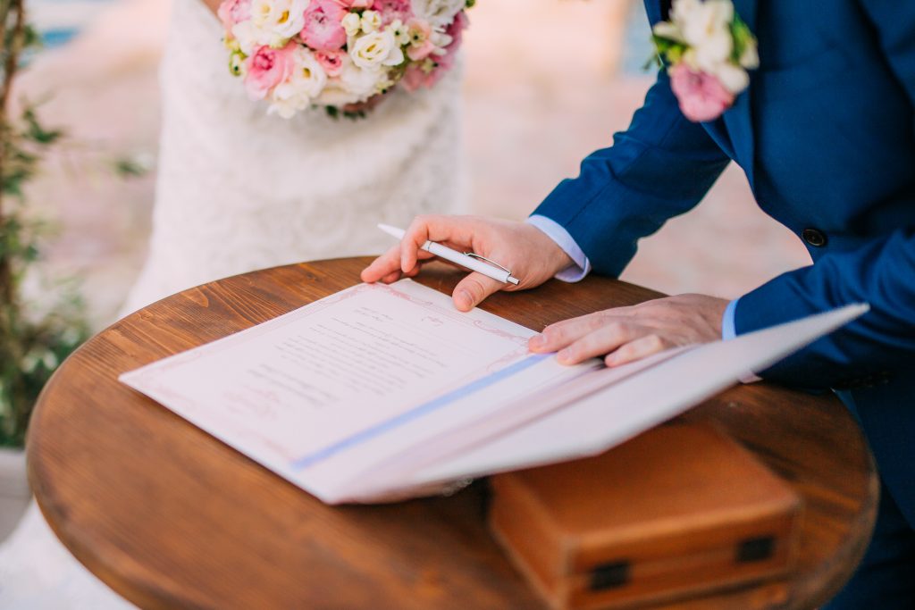 In questa foto sono inquadrate le mani di due sposi. Lo sposo sta firmando i documenti delle nozze civili su un tavolino di legno mentre la sposa lo guarda tenendo un bouquet di fiori rosa e bianchi