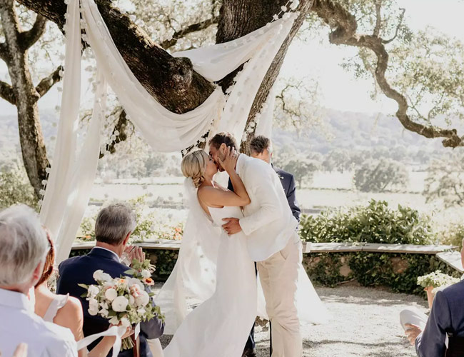 In questa foto due sposi si baciano al termine del loro matrimonio civile celebrato sotto una tenda di lino bianca in un campo di ulivi. Gli ospiti gli applaudono