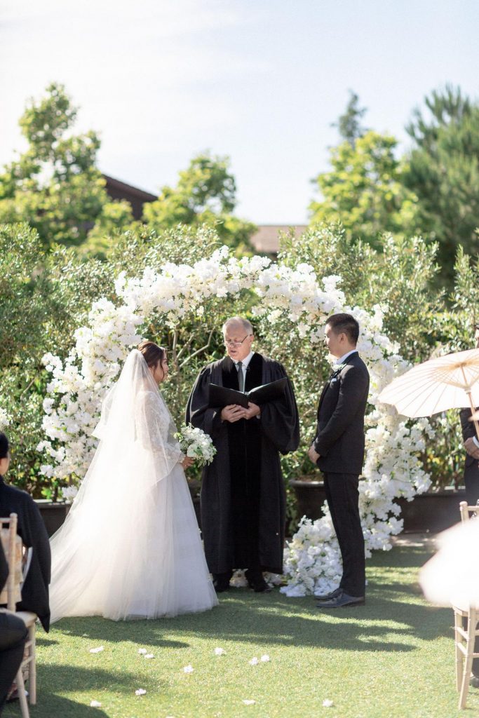 In questa foto l'officiante del matrimonio civile celebra il rito su un prato. Davanti a lui lo ascoltano i due sposi. Dietro di loro è stato allestito un cerchio di fiori bianchi 