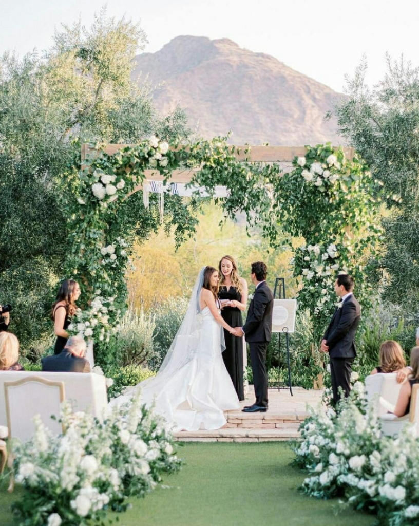 In questa foto un amica degli sposi celebra il loro matrimonio civile su un prato circondati da fiori e piante. Il rito si svolge sotto ad un gazebo di legno con piante rampicanti. Gli sposi si tengono per mano durante la celebrazione