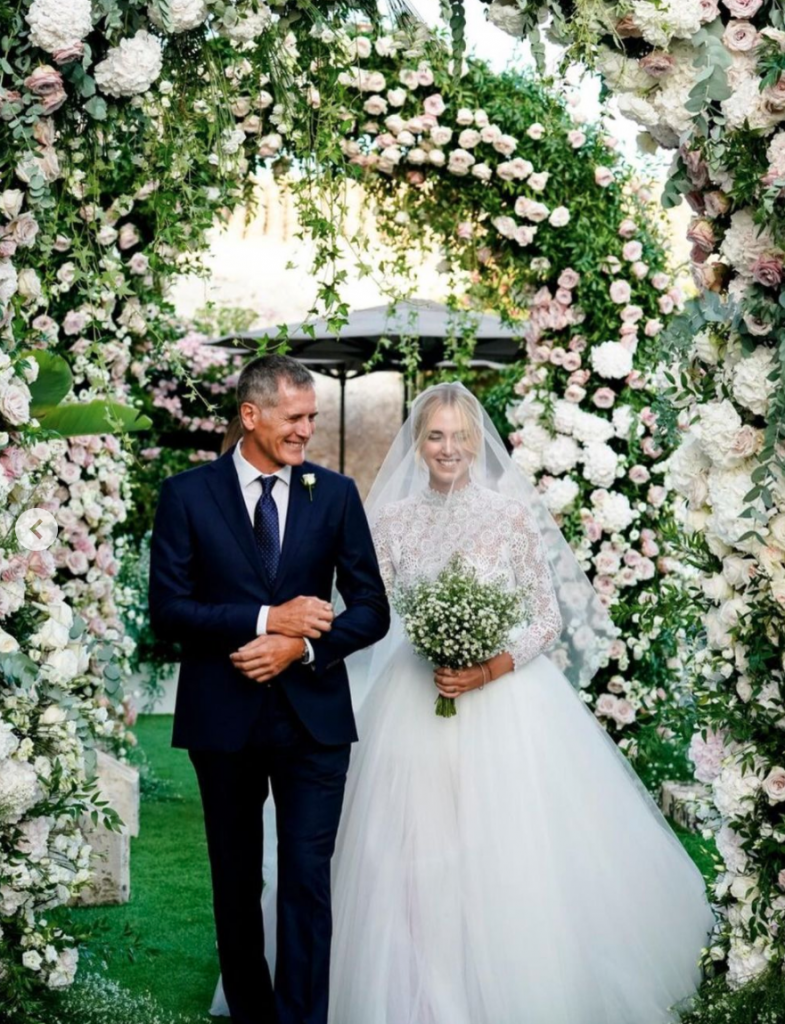 In questa foto Chiara Ferragni e il papà sorridenti fanno il loro ingresso nel giorno delle nozze. Sono circondati di fiori rosa e bianchi. Chiara Ferragni ha un bouquet di fiori velo di sposa