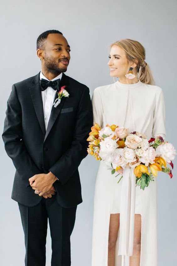 In questa foto due sposi si guardano sorridenti. Lei tiene un bouquet di fiori gialli e bianchi e indossa una tuta con shorts e giacca