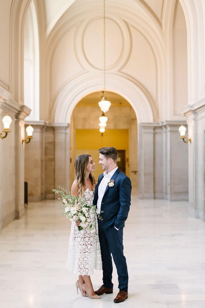 In questa foto due sposi si guardano sorridenti nel corridoio di marmo del Comune dove celebreranno il matrimonio. La sposa tiene nella mano destra un bouquet a braccio di fiori bianchi e rosa cipria. Lo sposo indossa un abito blu senza cravatta ma con bottoniera