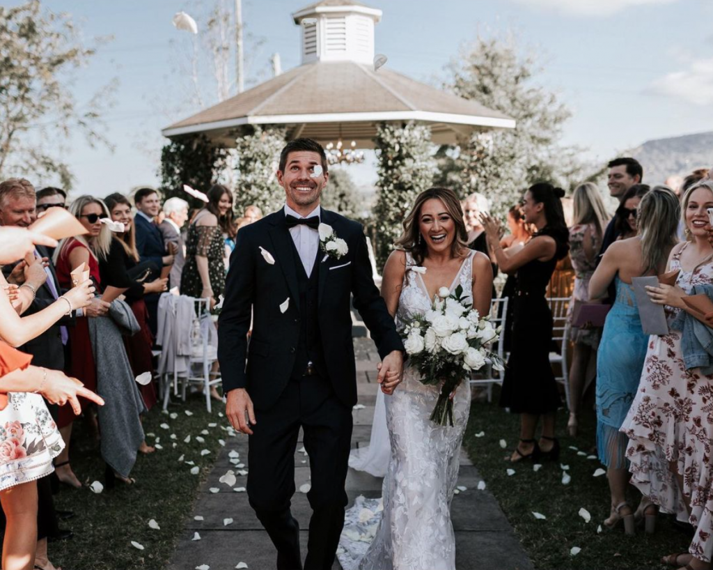 In questa foto due sposi escono sorridenti dalla location del rito civile in un prato con gazebo. Attorno a loro ci sono tutti gli invitati. Volano petali di rosa bianchi. La sposa indossa un abito a sirena con ricami floreali e tiene un boquet di fiori bianchi