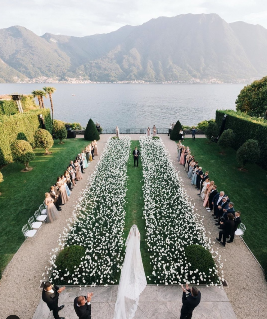 In questa foto l'ingresso della sposa nella location del suo matrimonio civile sul lago di Como lungo un corridoio di fiori bianchi disposti su un prato. Ai lati sono presenti gli invitati 