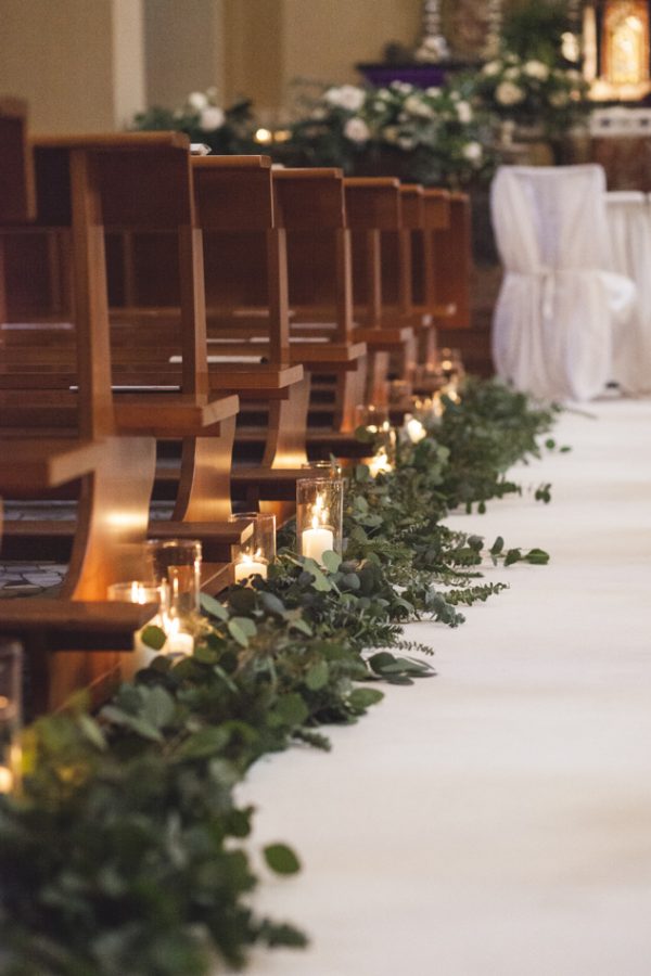 In questa foto una chiesa decorata con un tappeto bianco che ricopre la navata sui cui bordi sono sparse foglie di eucalipto. Sulle foglie sono poggiate candele accese dentro cilindri trasparenti. Sull'altare sono presenti composizioni di foglie e fiori bianchi. All'altare si intravedono sedie bianche