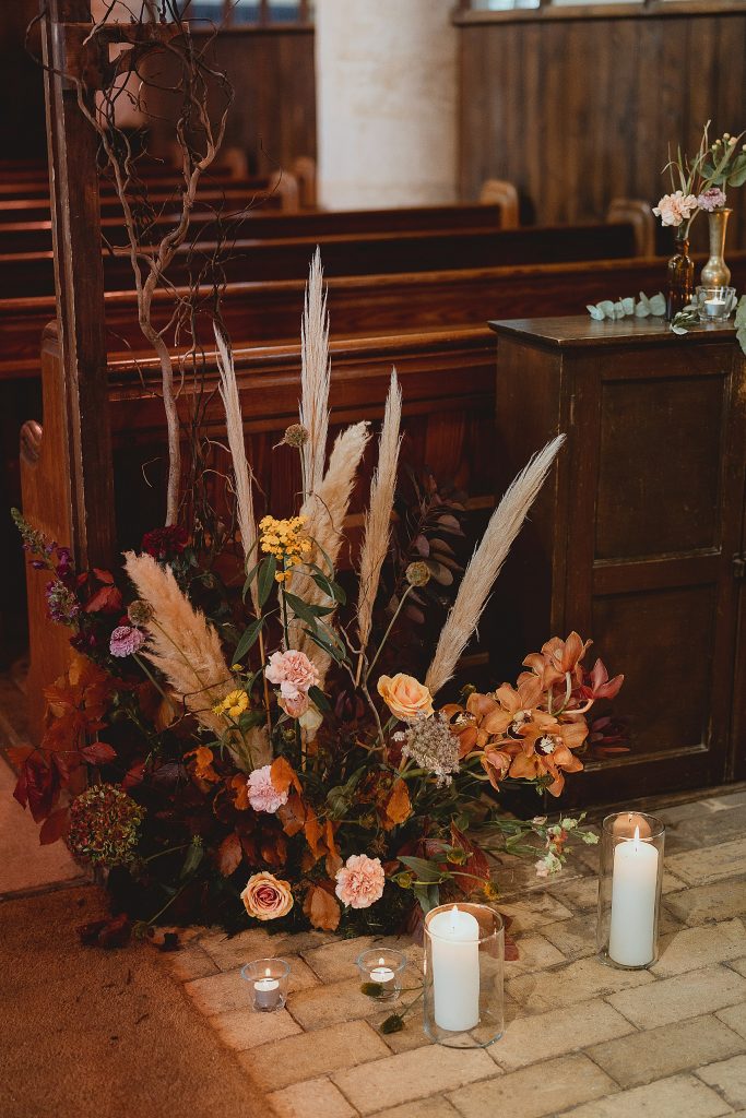 In questa foto una composizione floreale poggiata sul pavimento della chiesa in autunno realizzata con rose gialle, garofani rosa, orchidee arancioni, pampas, foglie e fiori bordeaux