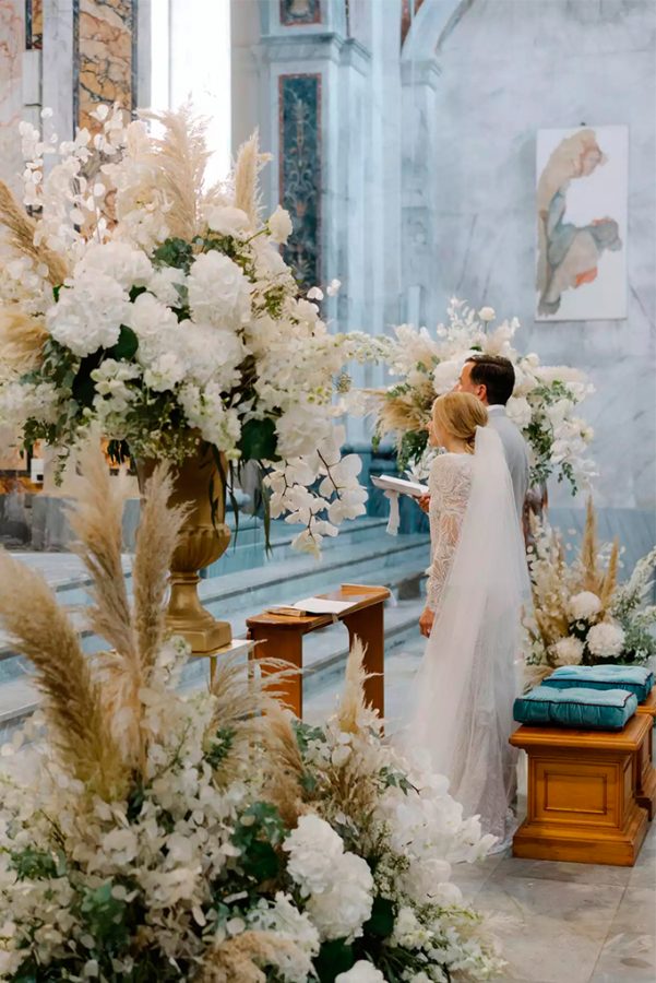 In questa foto due sposi in piedi all'altare ripresi di profilo. In primo piano vengono mostrati gli addobbi in stile shabby chic con pampas, fiori bianchi e foglie. Le stesse composizioni si intravedono sull'altro lato dell'altare. Sulle sedie di legno degli sposi sono poggiati 2 cuscini quadrati di colore turchese abbinati al marmo dei gradi presenti davanti all'altare. Davanti agli sposi è presente un inginocchiatoio su cui è poggiato il libretto messa della sposa