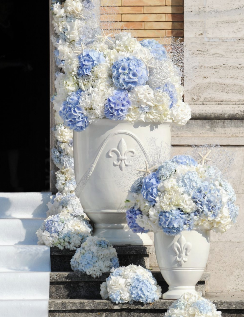 In questa foto addobbi per l'esterno di una chiesa per matrimonio realizzati con ortensie azzurre e bianche. Sui gradini dono sistemati due vasi con all'interno le composizioni di ortensie: il vaso in primo piano è più piccolo di quello più grande in secondo piano. Sui gradini accanto ai vasi sono poggiate piccole composizioni semisferiche di ortensie abbinate. Le ortensie circondano anche l'ingresso della chiesa. Sui gradini di marmo grigio si intravede disteso anche un tappeto bianco