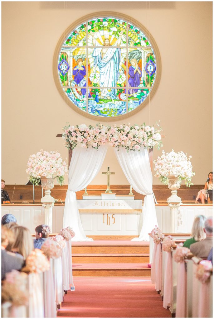 In questa foto una chiesa per matrimonio decorata con rose rosa e bianche lungo la navata e all'altare. Ai banchi della chiesa sono legati piccoli mazzi di fiori da cui pendono nastri rosa cipria. All'altare è montato un arco di legno con due drappi di stoffa bianca. La trave orizzontale è ricoperta di fiori. Ai lati dell'arco sono presenti due grandi vai bianchi poggiati du colonne con composizioni di fiori. Dietro l'arco, al centro è posizionato un crocifisso. Sull'altare è presente una vetrata colorata rotonda  che raffigura il Cristo pantocreatore